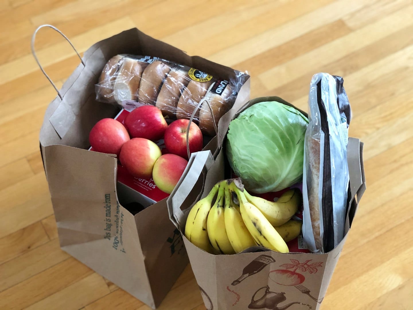 Zwei Einkaufstaschen voller frischem Obst, Gemüse und Backwaren auf einem Holzboden.