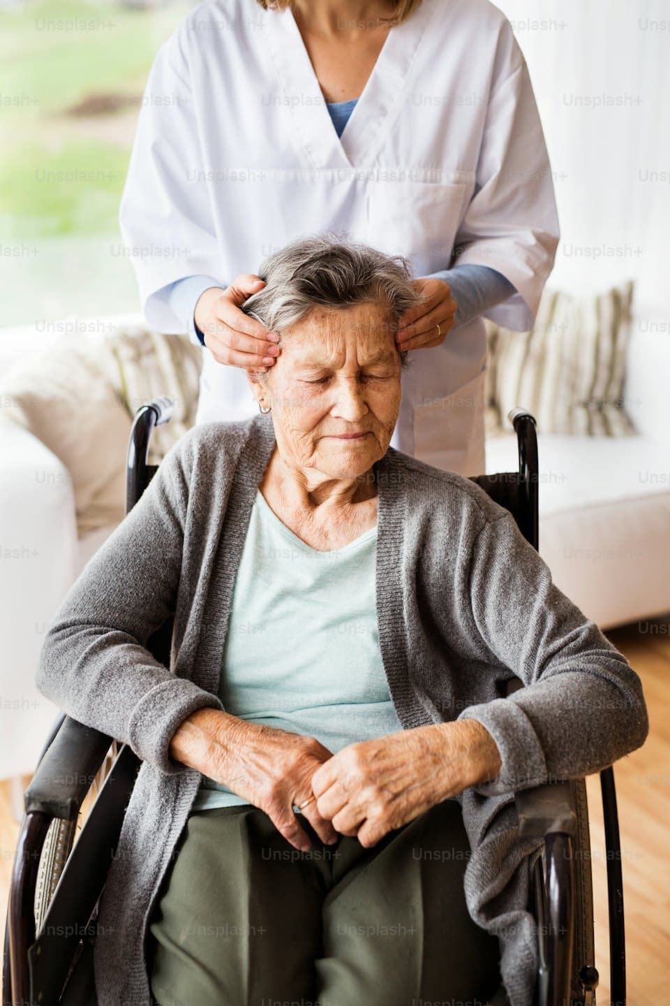 Pflegekraft massiert sanft den Kopf einer Frau im Rollstuhl.